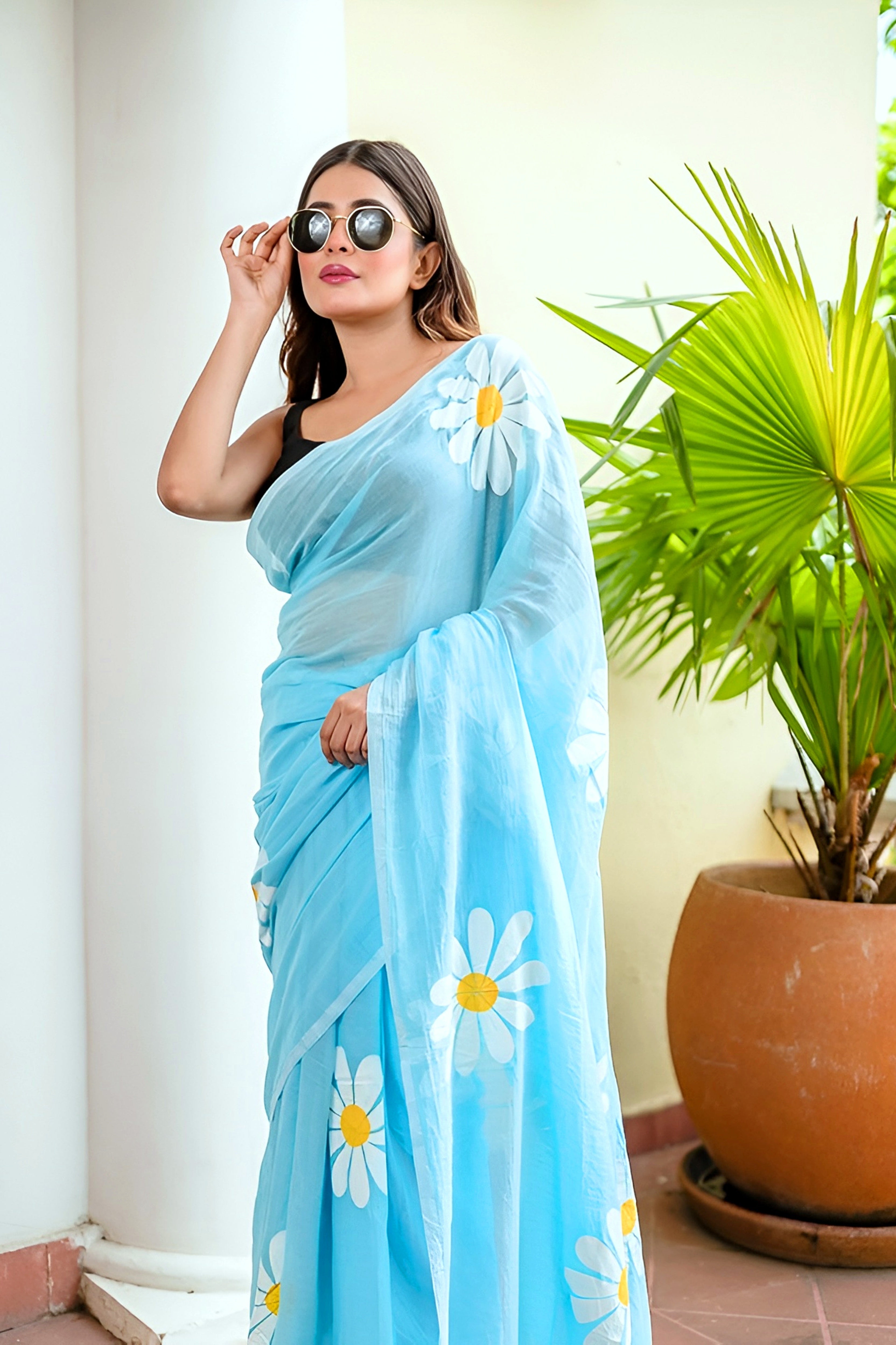 Woman wearing a light blue saree with white daisy patterns, standing next to a potted plant.