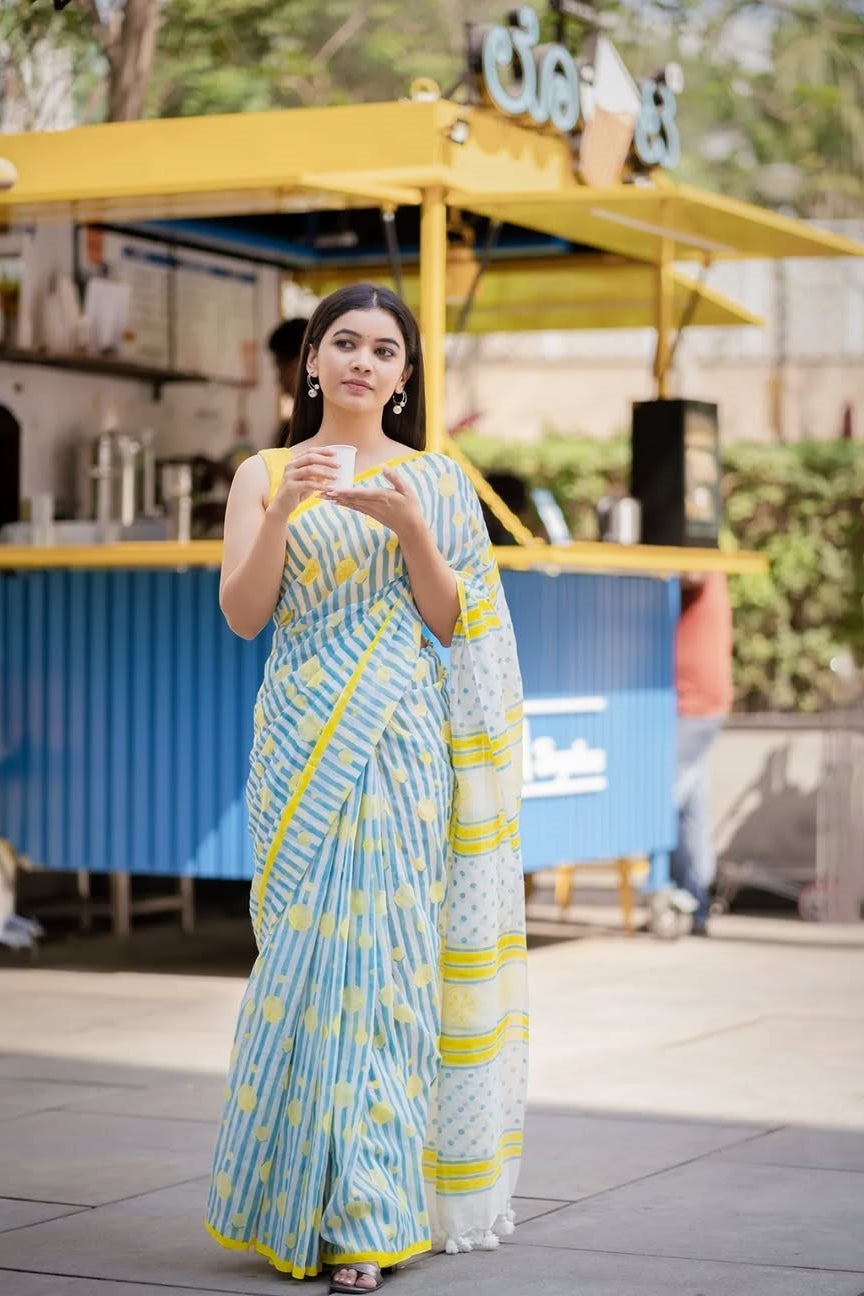 Woman in a blue and yellow saree holding a cup outdoors with a food truck in the background.