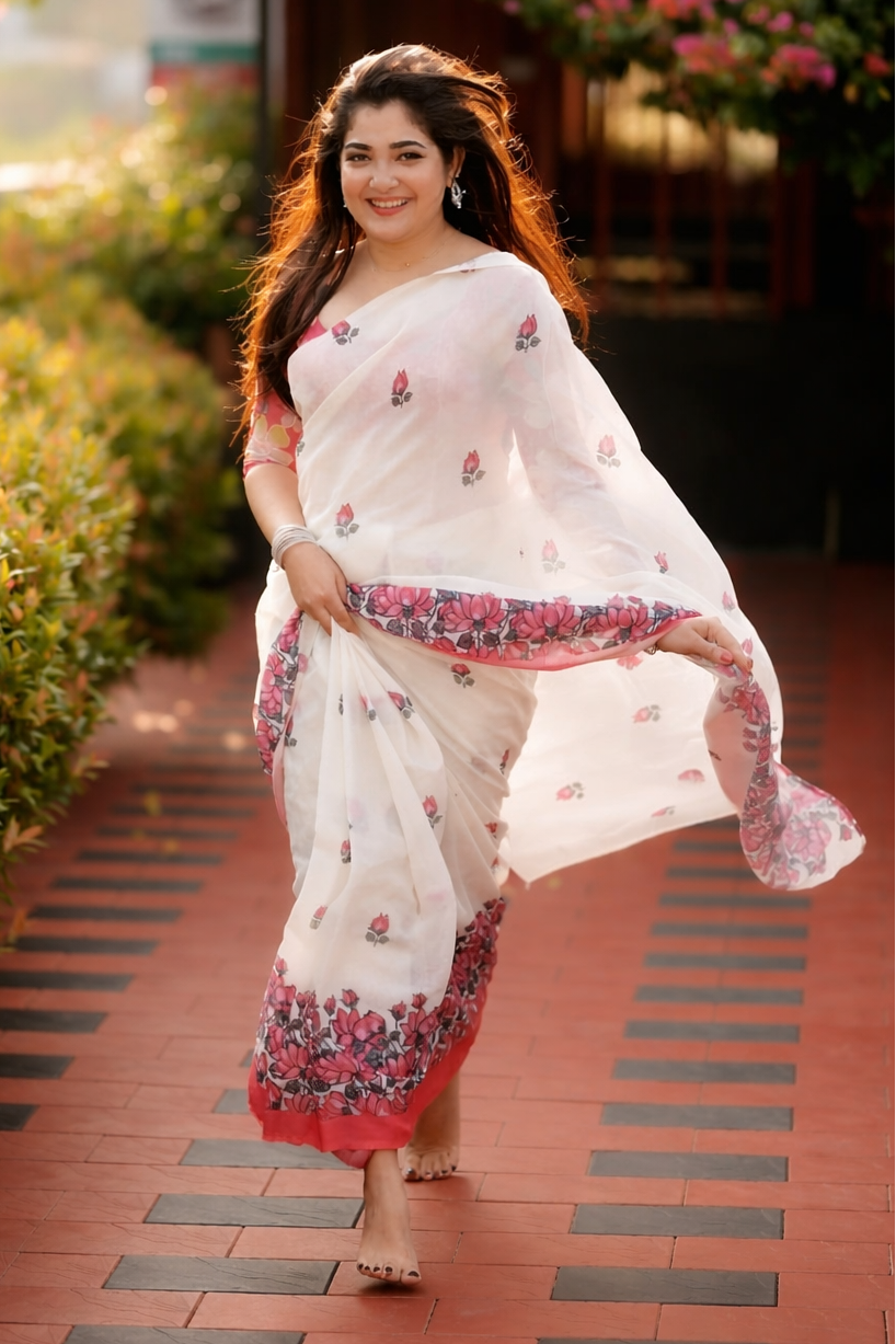 Woman in a white saree with red and pink patterns walking on a path with greenery around.