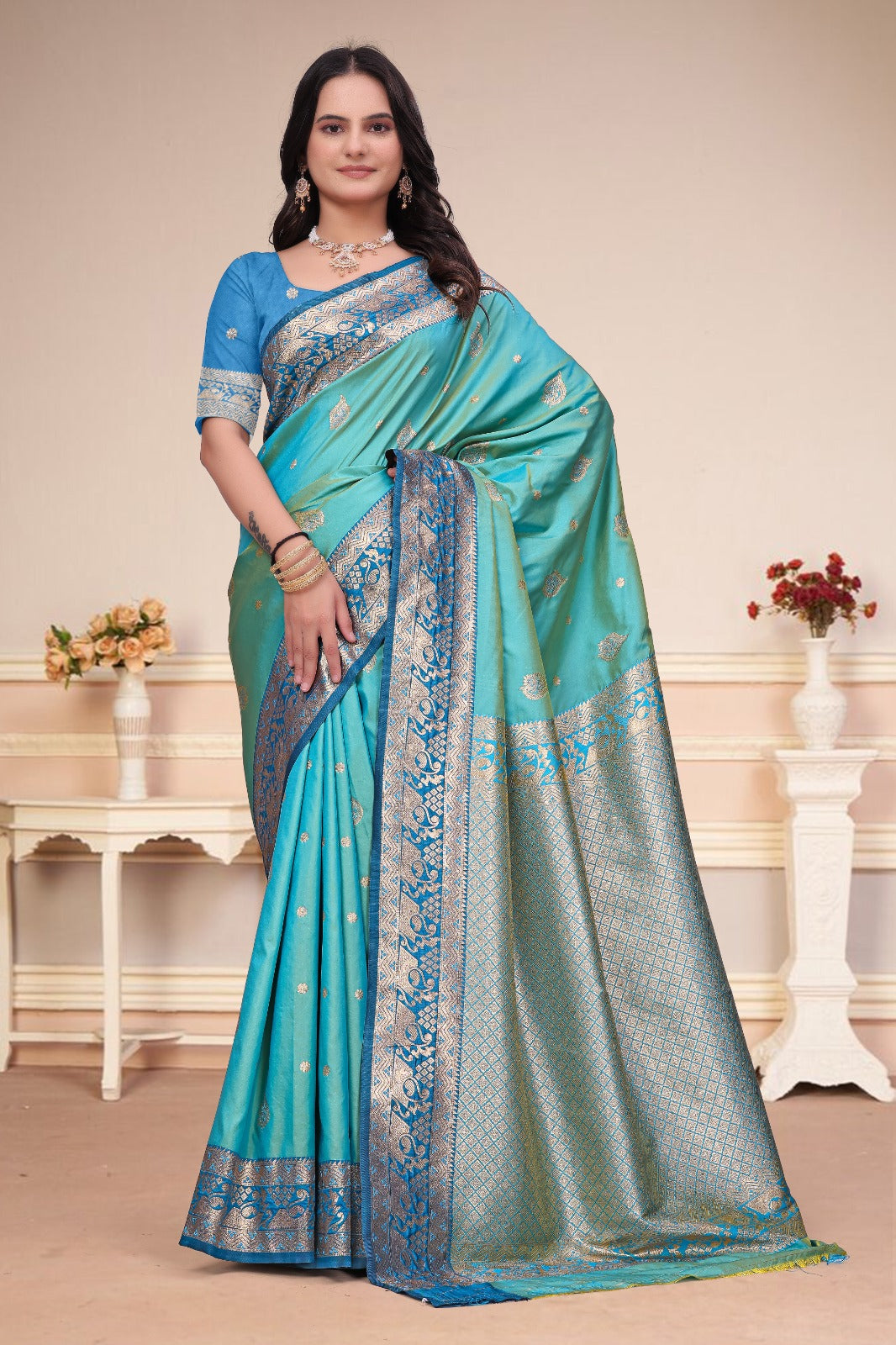 A woman modeling a turquoise blue and silver woven saree with a matching blue embroidered blouse, standing in a decorated indoor setting with cream walls and white side tables.