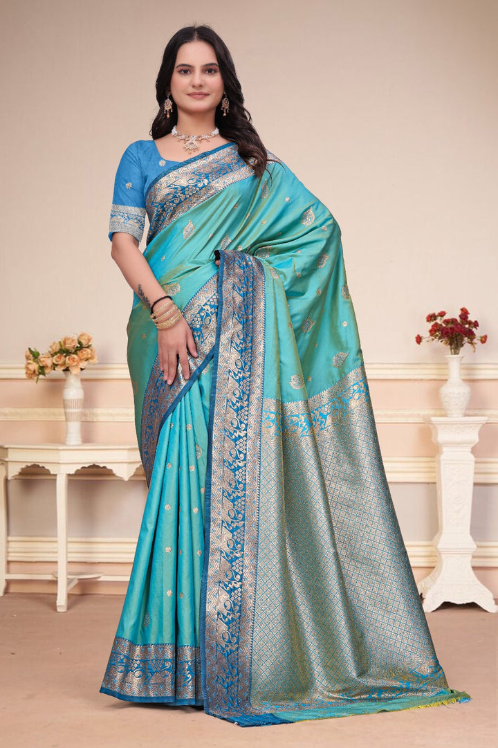 A woman modeling a turquoise blue and silver woven saree with a matching blue embroidered blouse, standing in a decorated indoor setting with cream walls and white side tables.