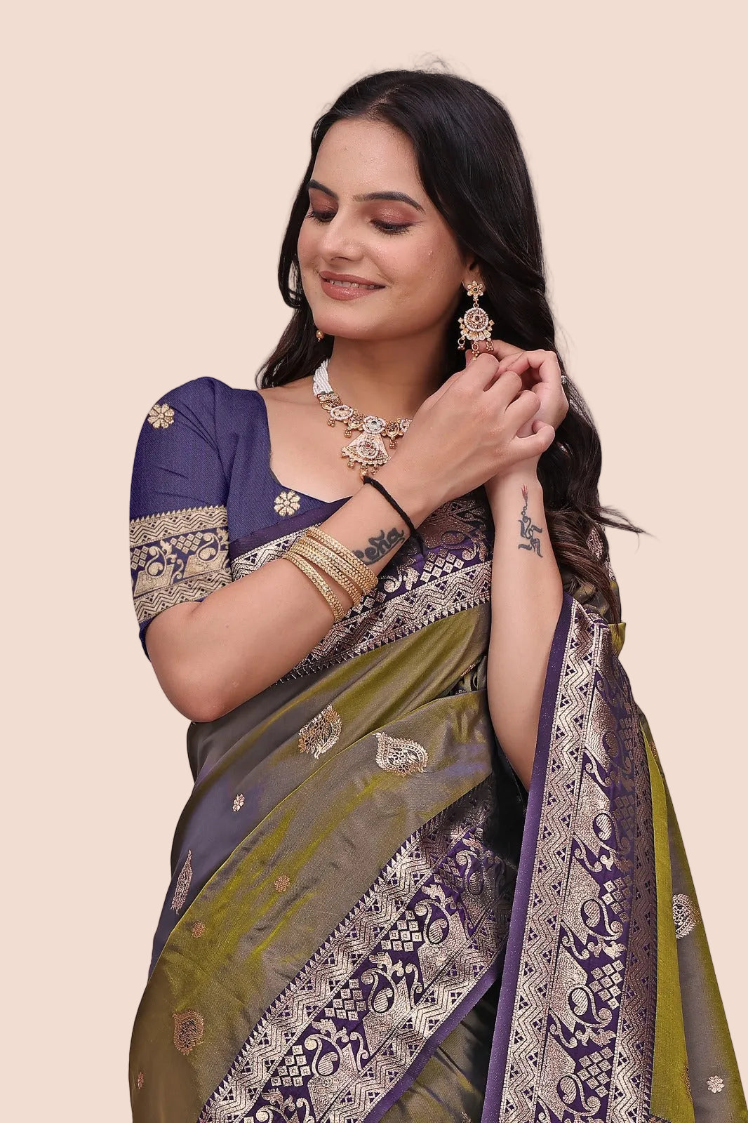 A woman wearing a purple and olive green Banarasi silk saree with intricate gold zari embroidery, paired with a purple embroidered blouse, gold jewelry, and bangles, posing against a neutral beige background.