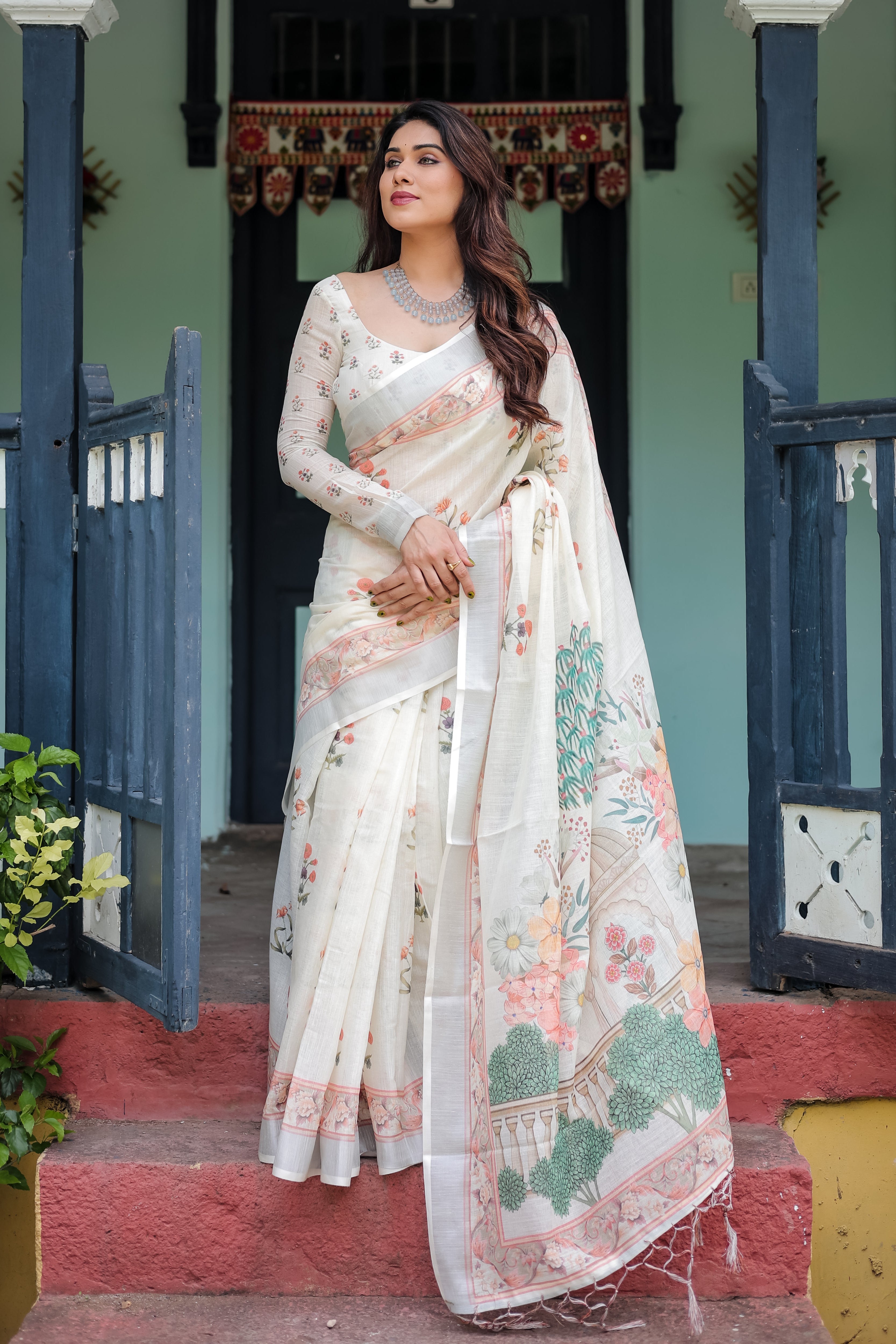 Woman in a white saree with floral patterns standing in front of a traditional building.