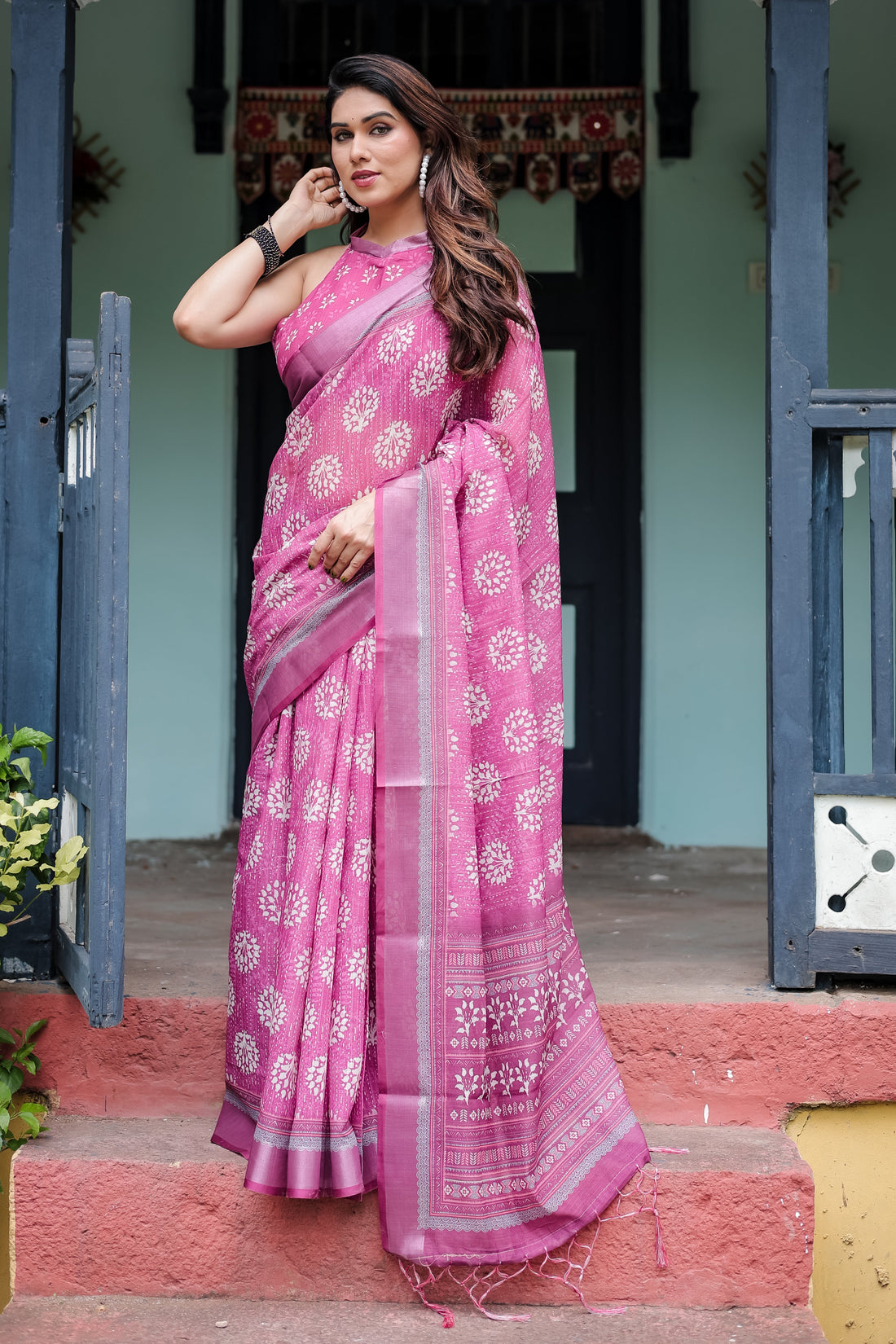 Woman in a pink saree standing in front of a traditional house entrance.