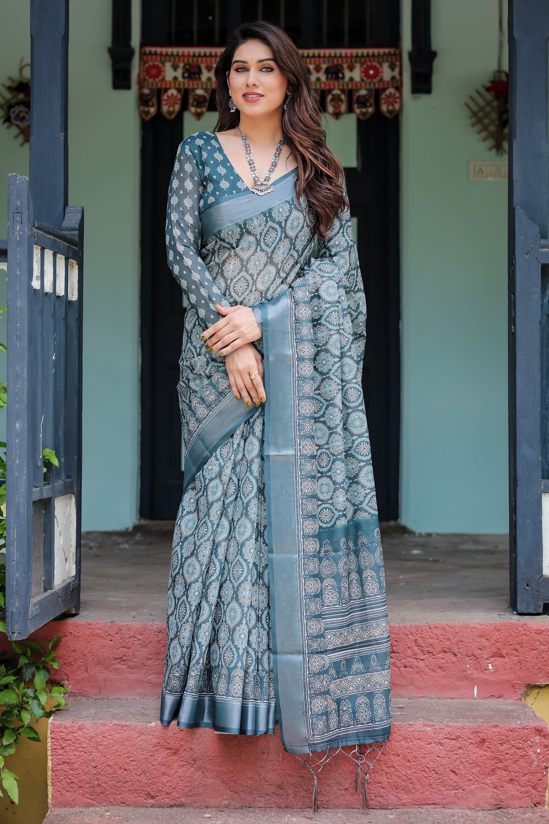 Woman in a blue saree standing on steps with a traditional door and wall in the background