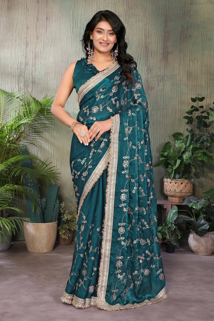 A woman modeling a teal green saree with intricate silver floral embroidery and a beige border, standing in front of a textured green wall with potted plants.