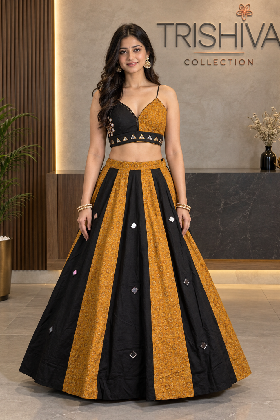 A woman models a black and mustard yellow traditional lehenga choli set in a boutique setting with 'TRISHIVA COLLECTION' on the wall behind her.