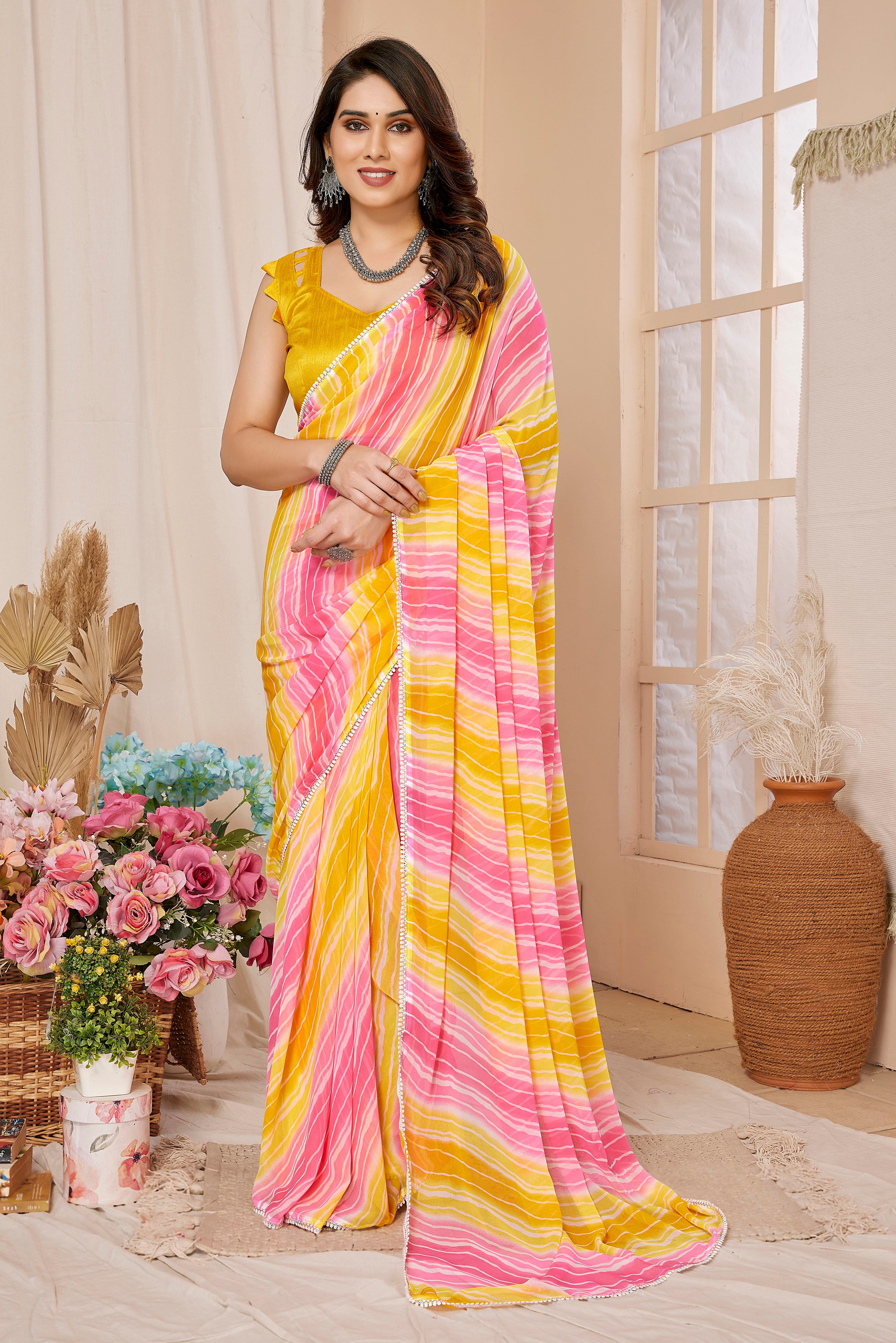 A woman models a bright yellow and pink striped georgette saree with a matching yellow sleeveless blouse, posed in a decorated indoor setting with flowers and a vase.