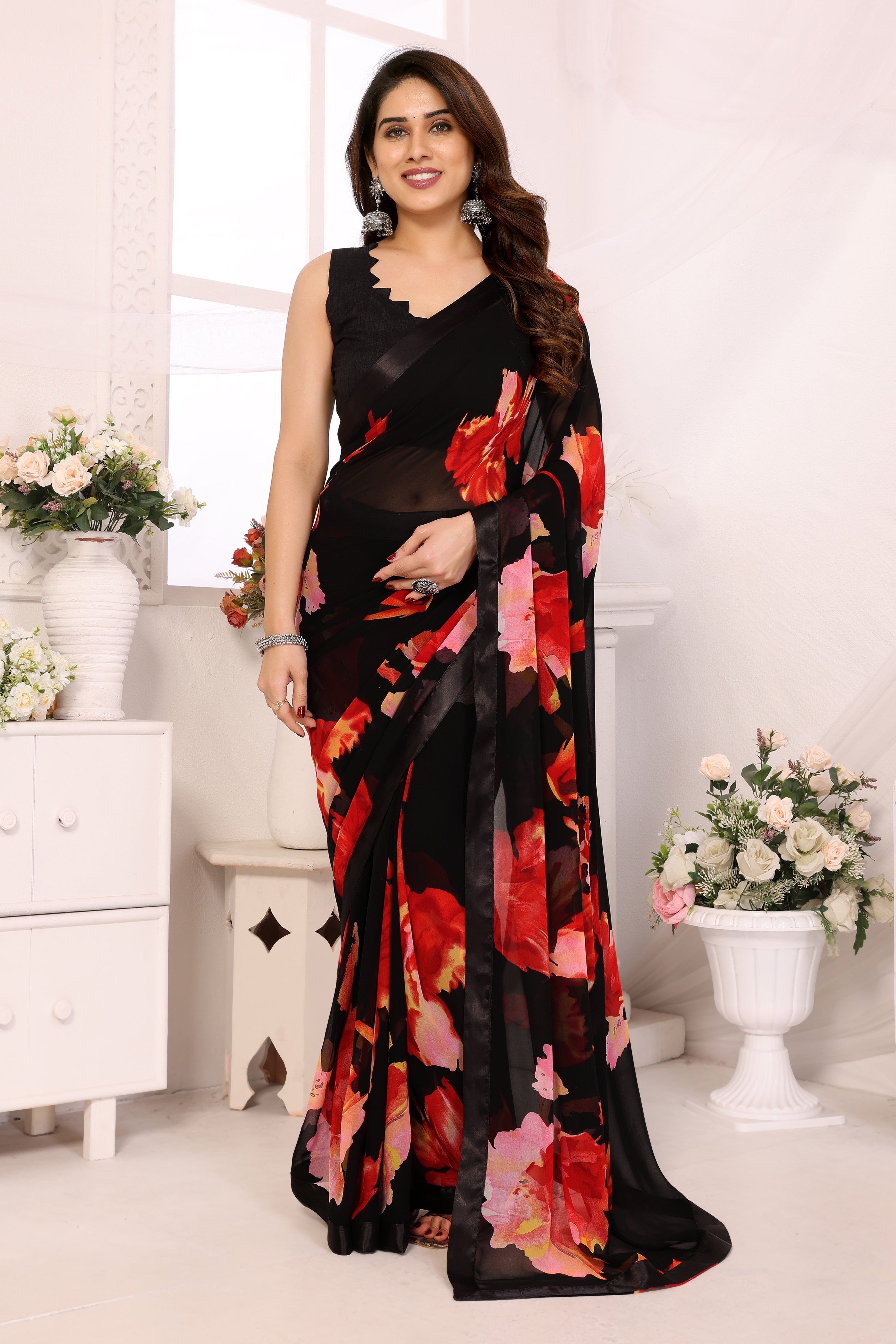 A woman modeling a black georgette saree with a vibrant red, pink, and orange floral print, paired with a matching black sleeveless blouse, in a bright white room decorated with flowers.
